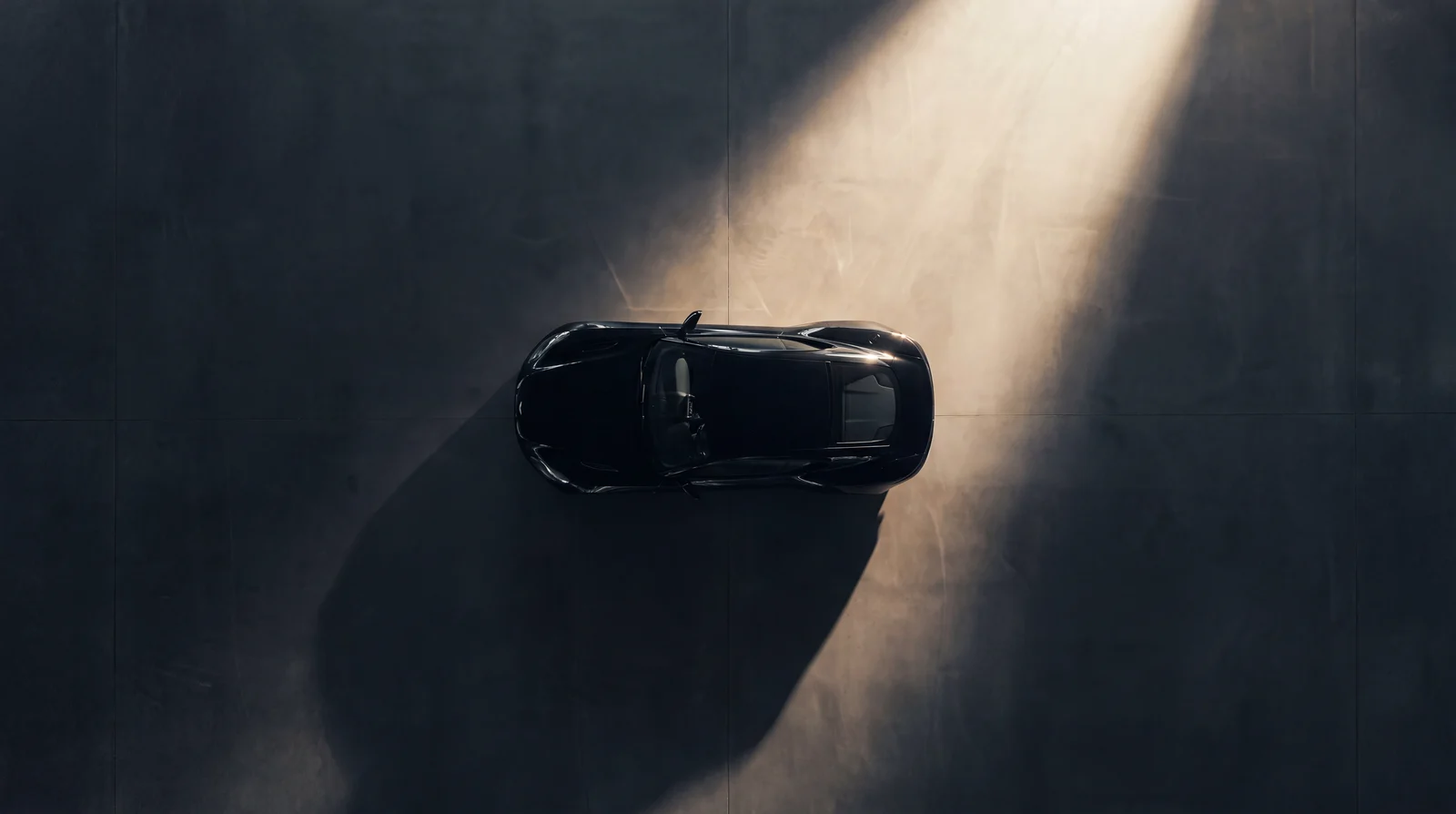 Overhead aerial view of a black coupé alone on a vast dark concrete plane.